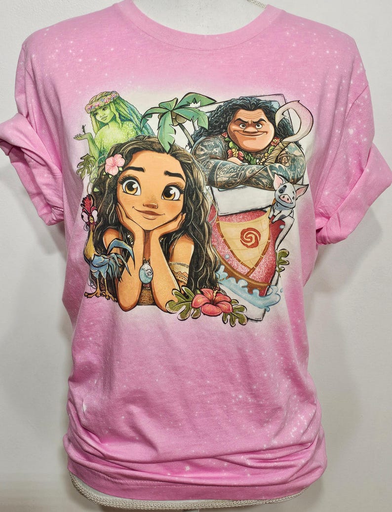 May include: Pink t-shirt featuring a graphic print of Moana and Maui from the Disney film Moana. The print also includes a green sea creature, a rooster, and a pig.