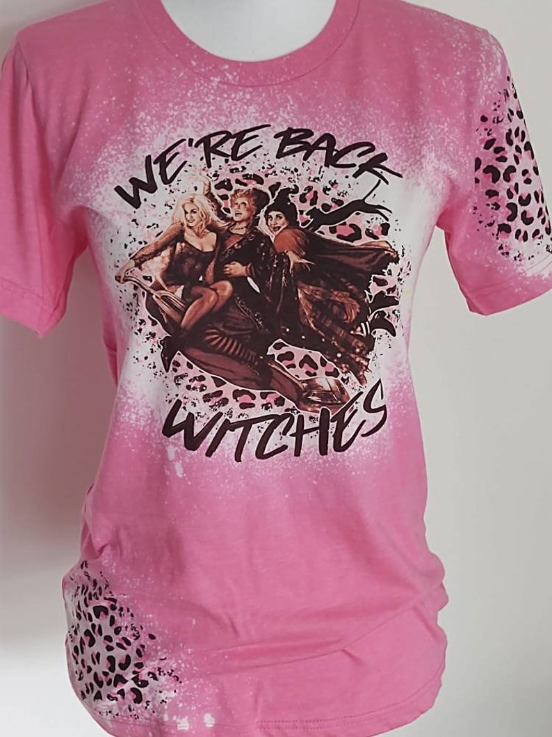 Puede incluir: Camiseta rosa con un dise&ntilde;o salpicado de lej&iacute;a que presenta a tres brujas volando en una escoba. El dise&ntilde;o incluye el texto "We're Back Witches" y un patr&oacute;n de estampado de leopardo.