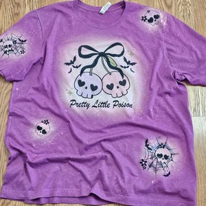 May include: Purple t-shirt with a distressed design featuring a pair of skull cherries with a black bow, bats, and the text "Pretty Little Poison." Additional skull and spiderweb graphics are on the sleeves and front.