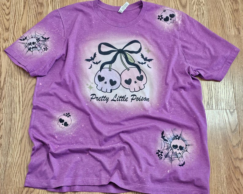 May include: Purple t-shirt with a distressed design featuring a pair of skull cherries with a black bow, bats, and the text "Pretty Little Poison." Additional skull and spiderweb graphics are on the sleeves and front.