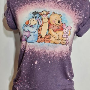 May include: A dusty purple short-sleeve t-shirt with a bleached effect. The center features a graphic illustration of Eeyore, Tigger, Winnie the Pooh, and Piglet sitting together. The fabric has a mottled, light-spotted appearance throughout.