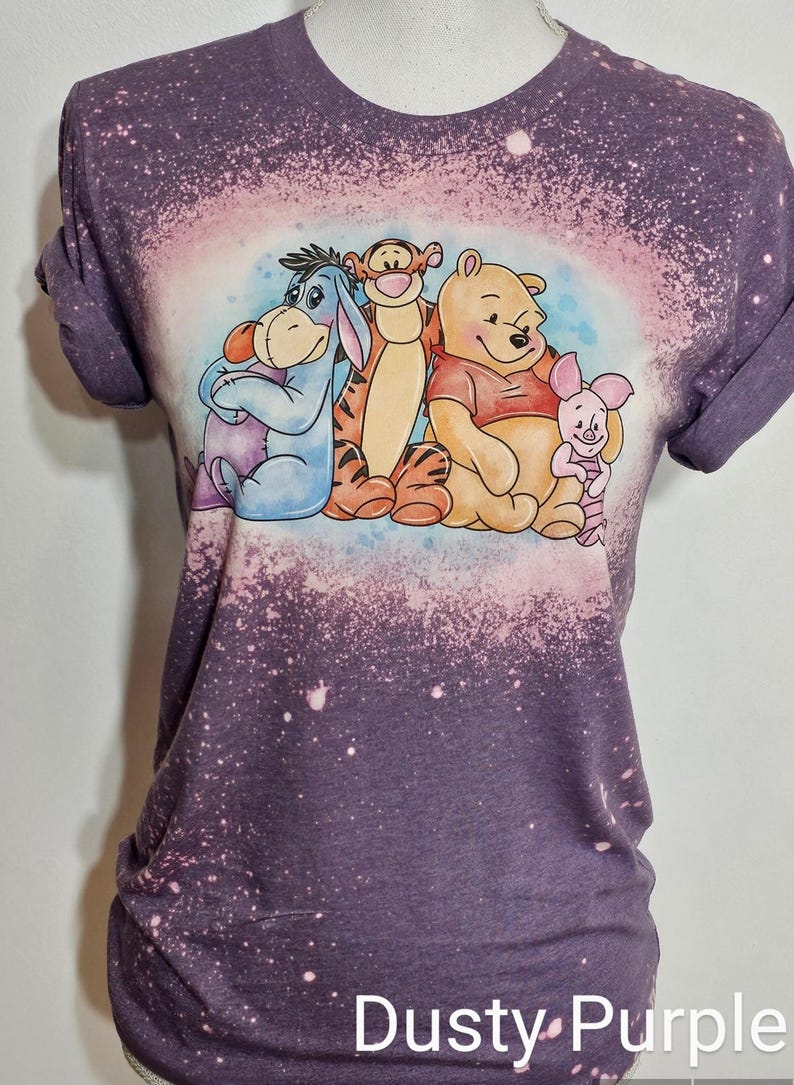 May include: A dusty purple short-sleeve t-shirt with a bleached effect. The center features a graphic illustration of Eeyore, Tigger, Winnie the Pooh, and Piglet sitting together. The fabric has a mottled, light-spotted appearance throughout.