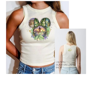 Peut inclure: D&eacute;bardeur cr&egrave;me avec un graphisme color&eacute; d'une princesse en robe verte, dans un design d'oreille de Mickey Mouse. Le dos du d&eacute;bardeur pr&eacute;sente une couronne et une signature. Le d&eacute;bardeur est port&eacute; avec un short en jean bleu.