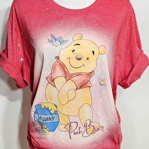May include: A red short-sleeved t-shirt with a bleached effect featuring a graphic of Winnie the Pooh, a blue bird, a butterfly, and a blue honey pot labeled "HUNNY". The text "Pooh Bear" is written in cursive script below the character.