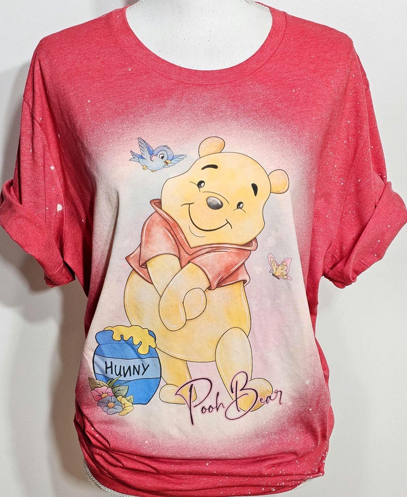 May include: A red short-sleeved t-shirt with a bleached effect featuring a graphic of Winnie the Pooh, a blue bird, a butterfly, and a blue honey pot labeled "HUNNY". The text "Pooh Bear" is written in cursive script below the character.