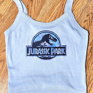 May include: A light blue vest top with a scoop neckline and thin shoulder straps. The centre of the garment features a circular graphic with a black silhouette of a dinosaur and the text JURASSIC PARK printed in a bold, stylised blue font.