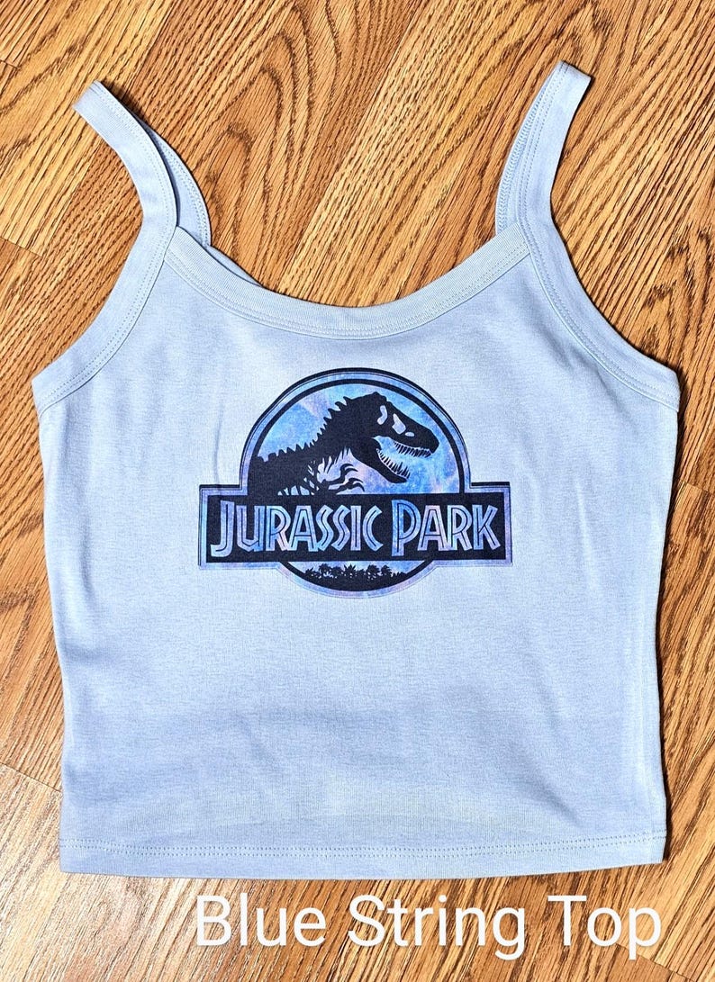 May include: A light blue vest top with a scoop neckline and thin shoulder straps. The centre of the garment features a circular graphic with a black silhouette of a dinosaur and the text JURASSIC PARK printed in a bold, stylised blue font.