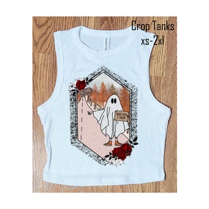 May include: White sleeveless crop tank top with a graphic print of a ghost holding a sign that says "Need Ride 2 Salem". The graphic is surrounded by red roses and a spider web.