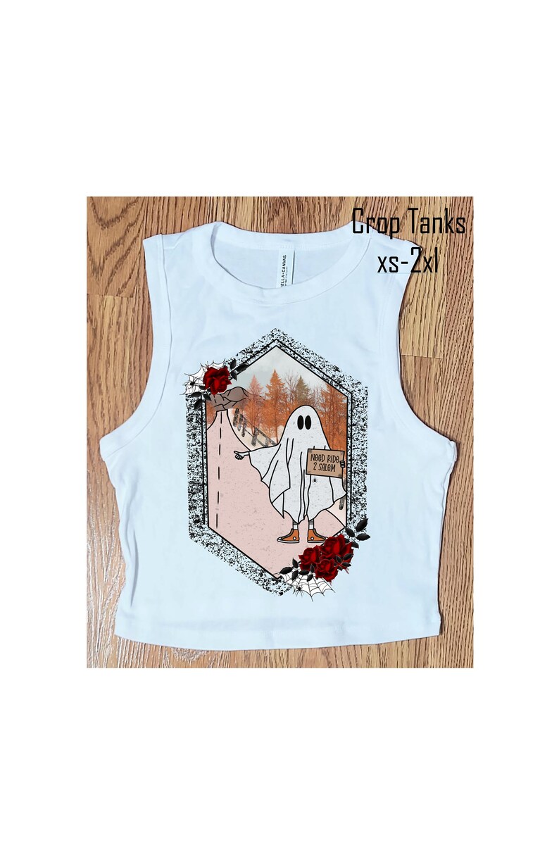 May include: White sleeveless crop tank top with a graphic print of a ghost holding a sign that says "Need Ride 2 Salem". The graphic is surrounded by red roses and a spider web.