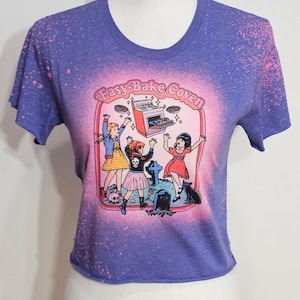 May include: A purple and pink tie-dye cropped t-shirt with a graphic of three girls in front of a stove with the text "Easy-Bake Coven".