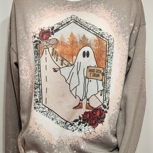 May include: A light brown sweatshirt with a bleached design of a ghost holding a sign that says "Need Ride 2 Salem". The ghost is wearing orange sneakers and is standing on a road with fall foliage in the background. The design is surrounded by a black hexagon with red roses and a spider web.