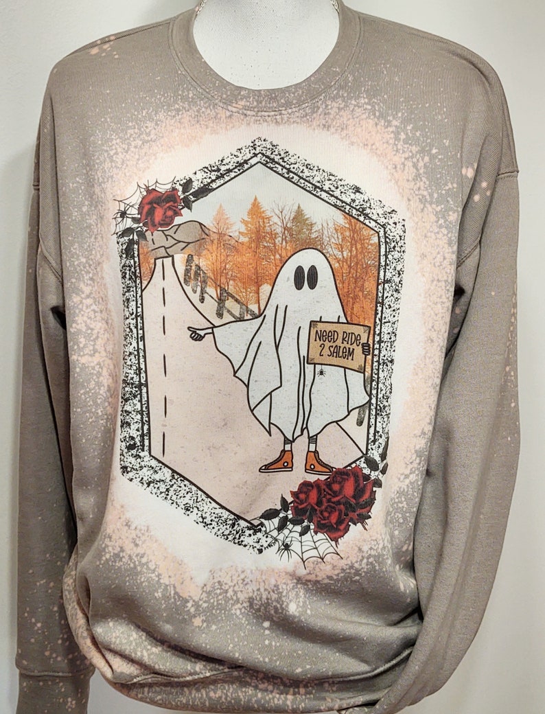 May include: A light brown sweatshirt with a bleached design of a ghost holding a sign that says "Need Ride 2 Salem". The ghost is wearing orange sneakers and is standing on a road with fall foliage in the background. The design is surrounded by a black hexagon with red roses and a spider web.
