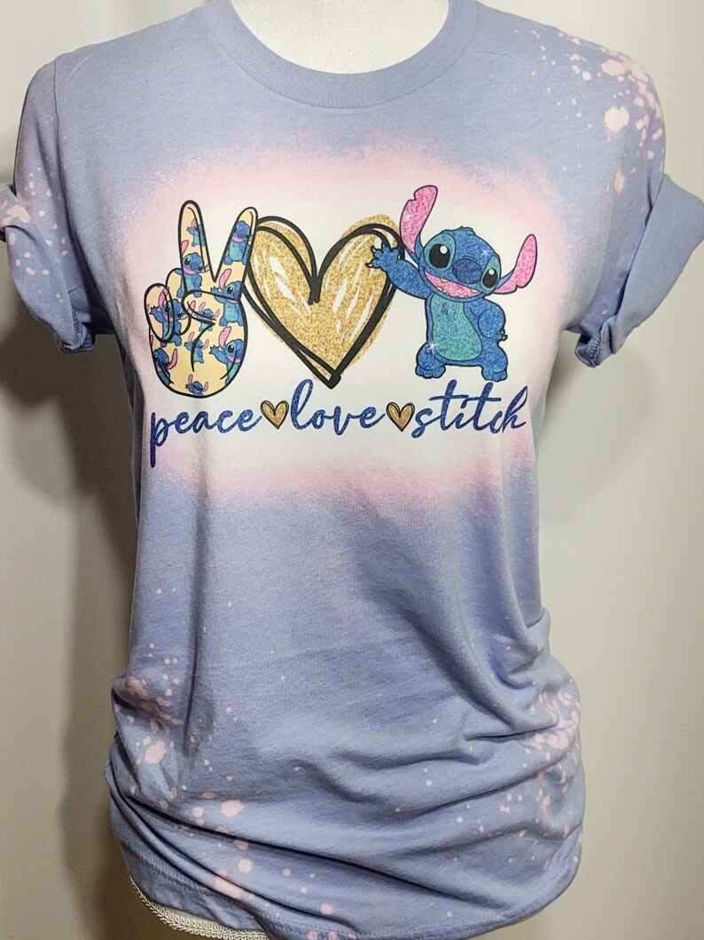 May include: A light blue t-shirt with a bleach-splattered design. The shirt features a peace sign, a heart, and a cartoon character with the text "peace love stitch".