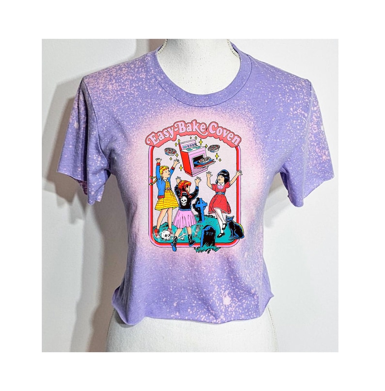 May include: Purple cropped t-shirt with a bleached effect. The shirt features a graphic of three girls in front of a tombstone with a flying Easy-Bake Oven and the text "Easy-Bake Coven".