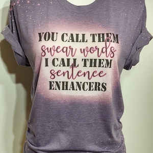 May include: A purple t-shirt with rolled-up sleeves and the text "YOU CALL THEM swear words I CALL THEM sentence ENHANCERS" in black and pink lettering. The shirt has a bleached, distressed look.