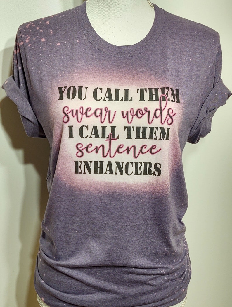 May include: A purple t-shirt with rolled-up sleeves and the text "YOU CALL THEM swear words I CALL THEM sentence ENHANCERS" in black and pink lettering. The shirt has a bleached, distressed look.