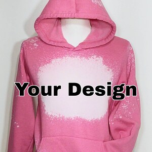 May include: Pink hoodie with a bleached white design area and a front pocket. The hoodie has a hood and long sleeves. The text "Your Design" is printed in black across the white area. The hoodie is displayed on a mannequin.
