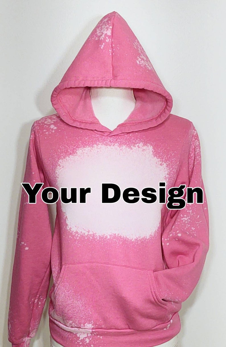 May include: Pink hoodie with a bleached white design area and a front pocket. The hoodie has a hood and long sleeves. The text "Your Design" is printed in black across the white area. The hoodie is displayed on a mannequin.