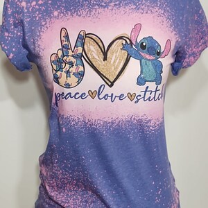 May include: A blue t-shirt with a pink and white bleach splatter design. The shirt features a peace sign made of Stitch characters, a gold heart, and the text "peace love stitch".