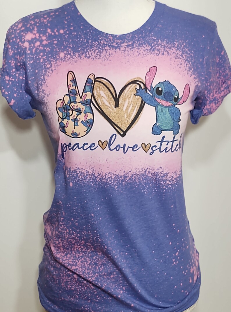 May include: A blue t-shirt with a pink and white bleach splatter design. The shirt features a peace sign made of Stitch characters, a gold heart, and the text "peace love stitch".