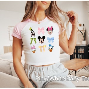 May include: Pink and white baby tee with a graphic print of Disney characters including Mickey Mouse, Minnie Mouse, Donald Duck, and Goofy. The characters are wearing bows and are surrounded by colorful bows.