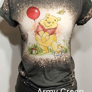 May include: An army green short-sleeve t-shirt with a bleached, mottled effect. The center features a graphic of a yellow bear wearing a red shirt, holding a red balloon, with a small bird and the handwritten text Pooh Bear below.
