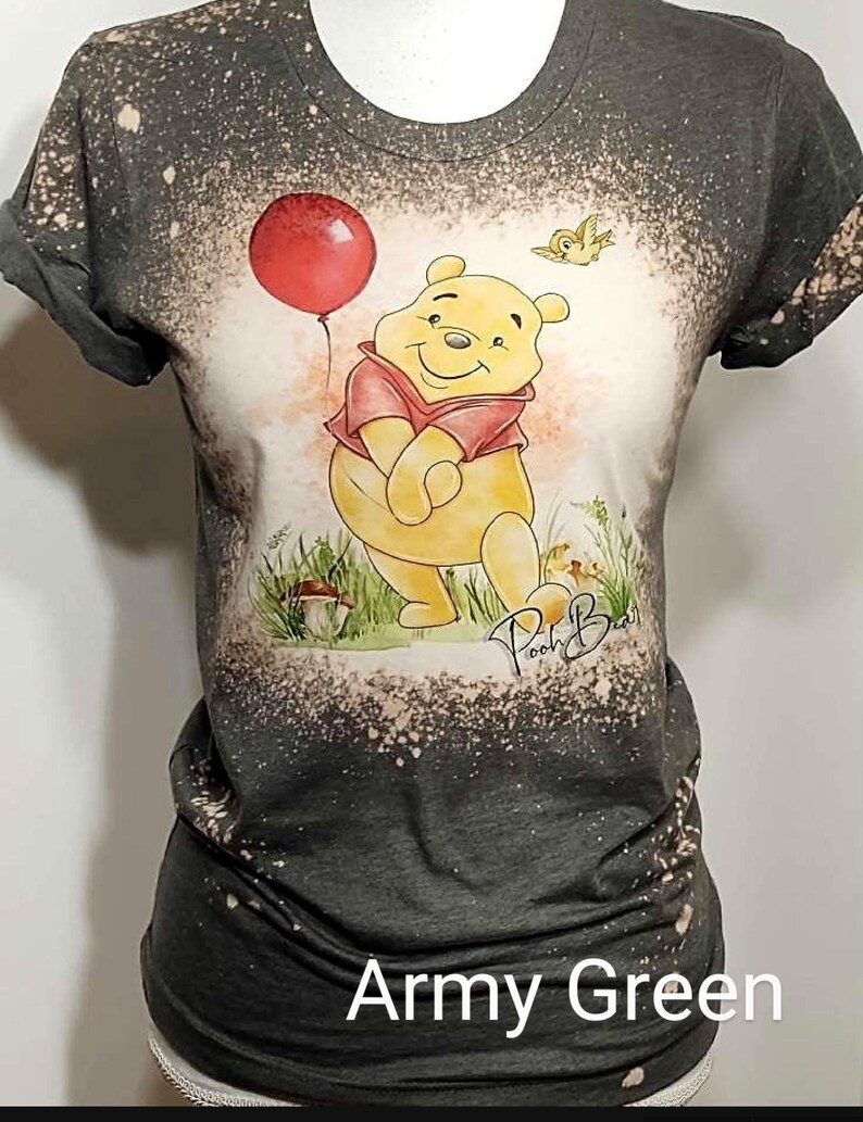 May include: An army green short-sleeve t-shirt with a bleached, mottled effect. The center features a graphic of a yellow bear wearing a red shirt, holding a red balloon, with a small bird and the handwritten text Pooh Bear below.