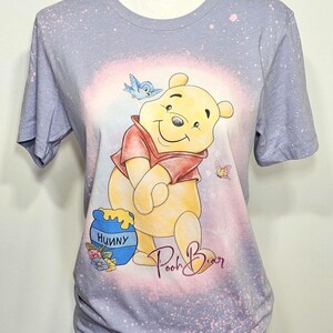 May include: A light purple short-sleeve t-shirt with a bleached effect featuring a graphic of Winnie the Pooh, a blue bird, a butterfly, and a blue honey pot labeled Hunny. The text Pooh Bear is printed in a cursive script below the character.