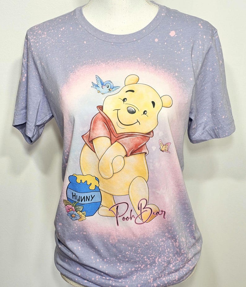 May include: A light purple short-sleeve t-shirt with a bleached effect featuring a graphic of Winnie the Pooh, a blue bird, a butterfly, and a blue honey pot labeled Hunny. The text Pooh Bear is printed in a cursive script below the character.