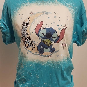 May include: A turquoise blue t-shirt with a white bleach design of Stitch from Lilo & Stitch holding Baby Yoda. The design includes a crescent moon and stars. The text "I love you to the moon & back" is written in black.