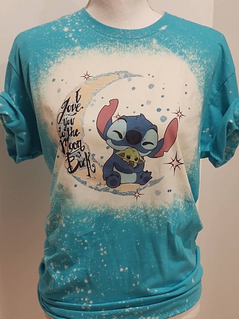 May include: A turquoise blue t-shirt with a white bleach design of Stitch from Lilo & Stitch holding Baby Yoda. The design includes a crescent moon and stars. The text "I love you to the moon & back" is written in black.