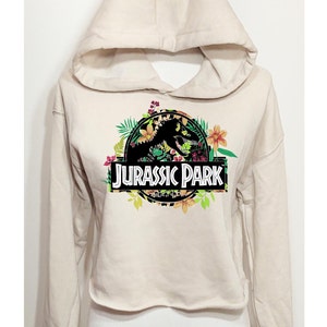 May include: A beige cropped hoodie featuring a graphic print of the Jurassic Park logo. The logo is surrounded by colourful tropical flowers and green leaves. The hoodie has long sleeves, a hood, and a raw-cut hemline at the waist.