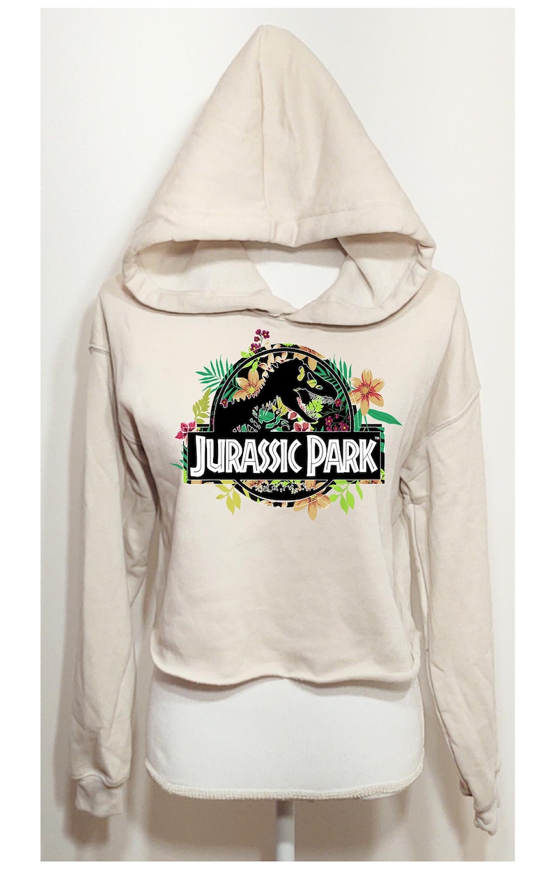 May include: A beige cropped hoodie featuring a graphic print of the Jurassic Park logo. The logo is surrounded by colourful tropical flowers and green leaves. The hoodie has long sleeves, a hood, and a raw-cut hemline at the waist.