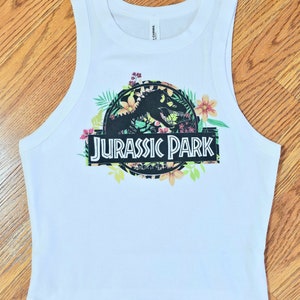 May include: A white sleeveless tank top featuring a graphic print of the Jurassic Park logo. The logo is surrounded by colourful tropical flowers and green leaves. The text JURASSIC PARK is printed in white letters on a black rectangular background.