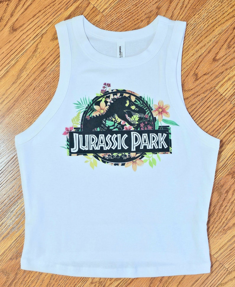 May include: A white sleeveless tank top featuring a graphic print of the Jurassic Park logo. The logo is surrounded by colourful tropical flowers and green leaves. The text JURASSIC PARK is printed in white letters on a black rectangular background.