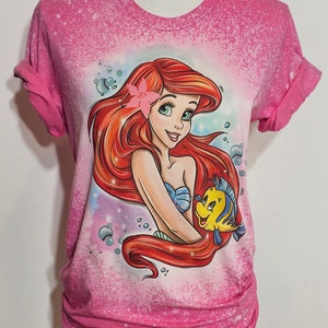 Puede incluir: Camiseta rosa con un dise&ntilde;o salpicado de lej&iacute;a que presenta una imagen de dibujos animados de Ariel de La Sirenita con Flounder.