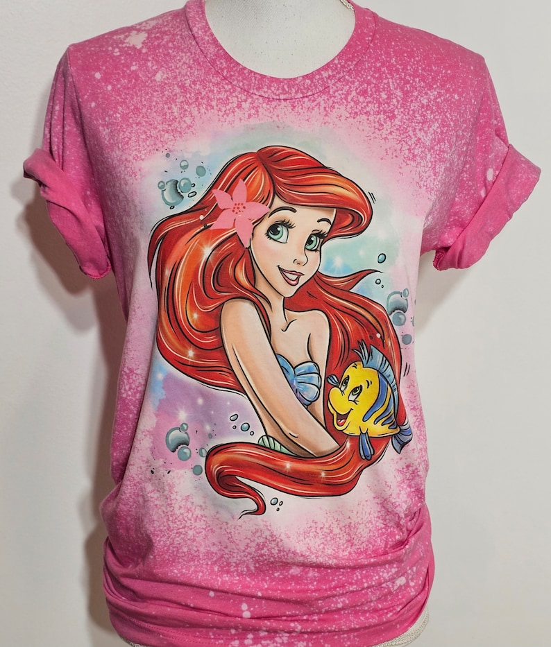 Puede incluir: Camiseta rosa con un dise&ntilde;o salpicado de lej&iacute;a que presenta una imagen de dibujos animados de Ariel de La Sirenita con Flounder.