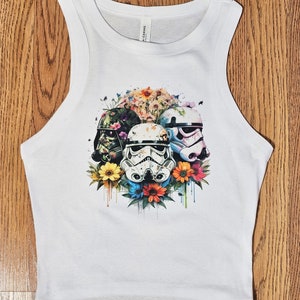 May include: White tank top with a graphic print of four Star Wars characters surrounded by colorful flowers. The characters are Darth Vader, a Stormtrooper, and two more Stormtroopers.