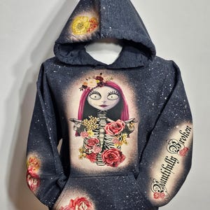 以下が含まれることがあります： 白い花とピンクの花の模様が施された濃いグレーのパーカー。花冠をかぶった骸骨と袖に「Beautifully Broken」の文字が描かれている。