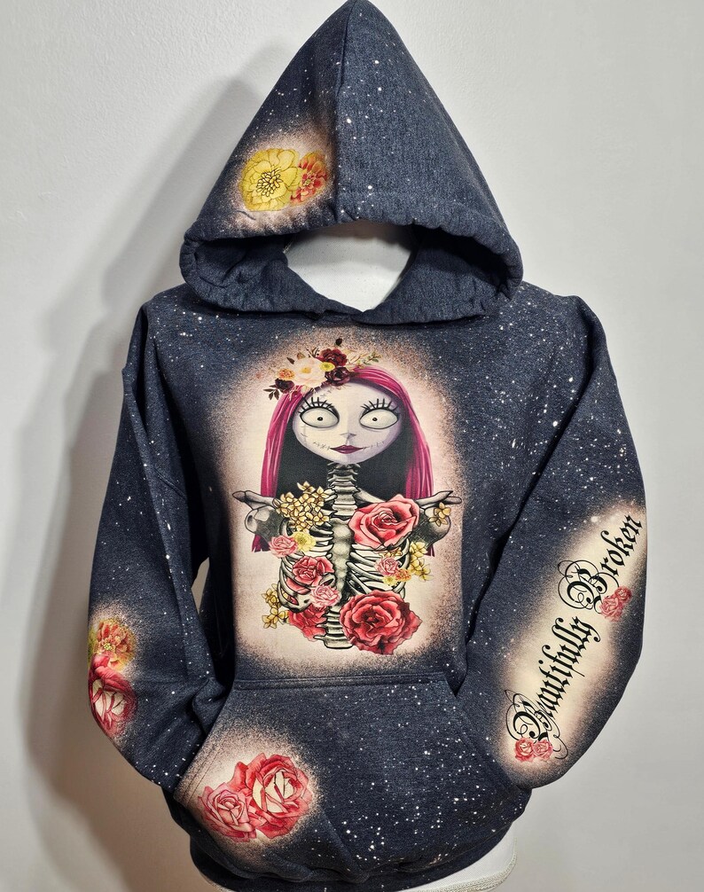 以下が含まれることがあります： 白い花とピンクの花の模様が施された濃いグレーのパーカー。花冠をかぶった骸骨と袖に「Beautifully Broken」の文字が描かれている。