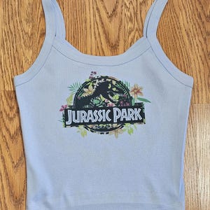 May include: A light blue vest top featuring a circular graphic with a black dinosaur silhouette, colourful tropical flowers, and green leaves. The text JURASSIC PARK is printed in white capital letters on a black rectangular banner across the centre.