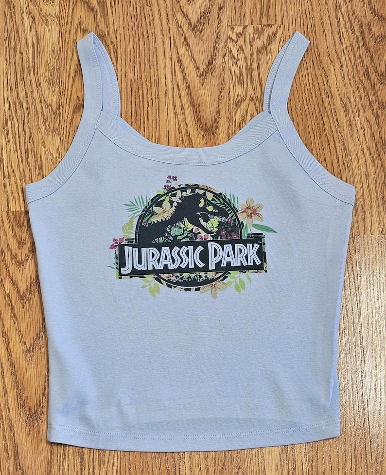 May include: A light blue vest top featuring a circular graphic with a black dinosaur silhouette, colourful tropical flowers, and green leaves. The text JURASSIC PARK is printed in white capital letters on a black rectangular banner across the centre.