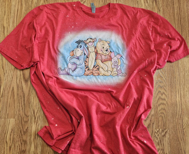 May include: A red short-sleeved t-shirt with a bleached-effect finish featuring a central graphic print of Eeyore, Tigger, Winnie the Pooh, and Piglet sitting together against a soft blue circular background on the front of the garment.
