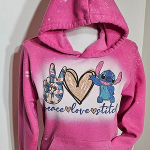 May include: Pink hoodie with a bleach-splattered design featuring a peace sign, a heart with glitter, and Stitch from Lilo & Stitch. The text "peace love stitch" is printed below the design.