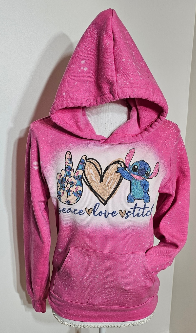 May include: Pink hoodie with a bleach-splattered design featuring a peace sign, a heart with glitter, and Stitch from Lilo & Stitch. The text "peace love stitch" is printed below the design.