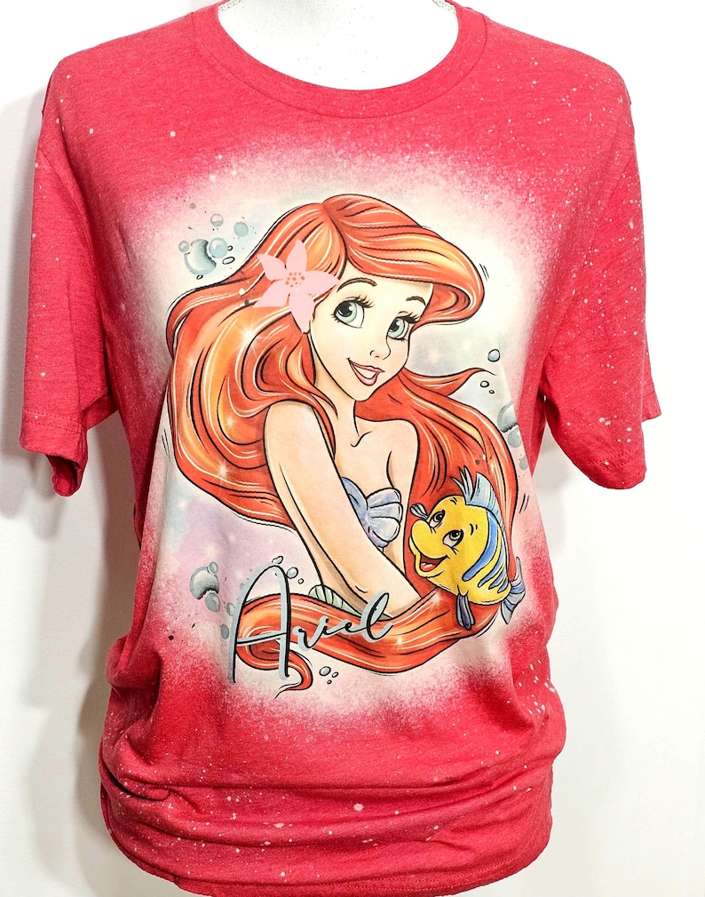 Puede incluir: Camiseta roja con un dise&ntilde;o salpicado de lej&iacute;a que presenta a Ariel de La Sirenita. Ariel est&aacute; sosteniendo a Flounder, un pez amarillo. La camiseta tiene la palabra "Ariel" escrita en cursiva en la parte inferior.