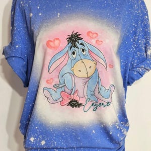 May include: A blue short-sleeved t-shirt with a bleached splatter pattern. The center features a graphic of the character Eeyore sitting among pink hearts, with a pink bow near the tail and the name Eeyore written in cursive script below.