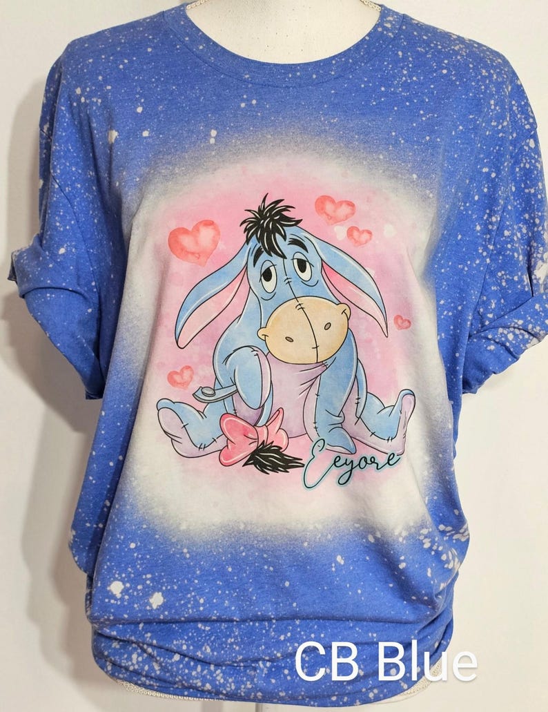 May include: A blue short-sleeved t-shirt with a bleached splatter pattern. The center features a graphic of the character Eeyore sitting among pink hearts, with a pink bow near the tail and the name Eeyore written in cursive script below.
