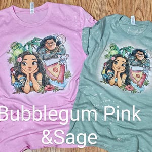 May include: Two t-shirts with a tie-dye effect. The pink shirt features a cartoon image of Moana and Maui from the Disney movie Moana. The green shirt features a cartoon image of Moana and Maui from the Disney movie Moana. The text "Bubblegum Pink & Sage" is printed on the bottom of the shirts.