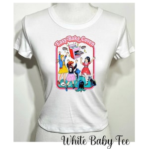 May include: White baby tee shirt with a graphic print of three girls in front of a pink Easy-Bake Oven. The girls are wearing colorful clothing and are holding up the oven. The graphic is surrounded by a red border and the text "Easy-Bake Coven" is written in pink letters.